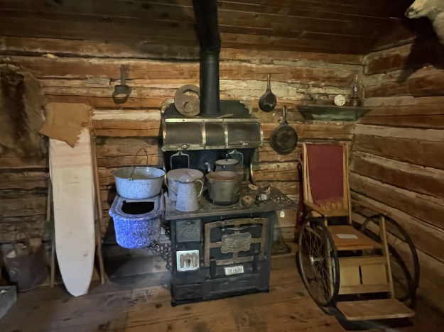 A frontier cabin showing the stove, utensils, ironing board and wheel chair of that time.